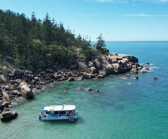 A white tour boat with blue accents floats in crystal clear turquoise water near a rocky, tree-covered island coastline under a bright blue sky.