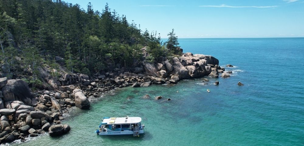 A white tour boat with blue accents floats in crystal clear turquoise water near a rocky, tree-covered island coastline under a bright blue sky.