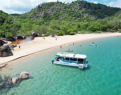 Catamaran anchored in clear turquoise water near a sandy beach lined with lush green hills and large rocks.