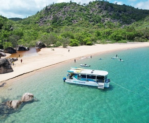 Catamaran anchored in clear turquoise water near a sandy beach lined with lush green hills and large rocks.