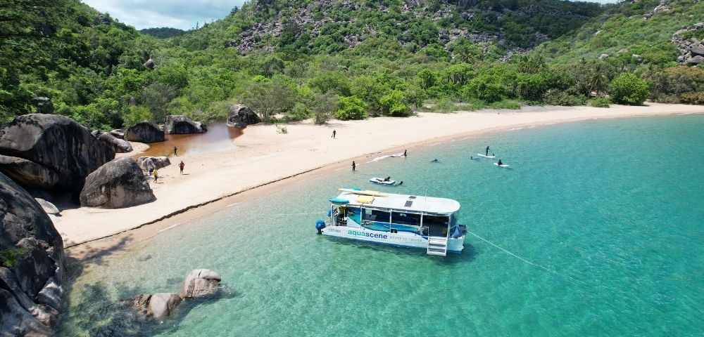 Catamaran anchored in clear turquoise water near a sandy beach lined with lush green hills and large rocks.