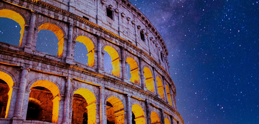 Illuminated Colosseum arches with a starry night sky backdrop. Warm yellow lights create a historic, serene ambiance against the vast cosmos.