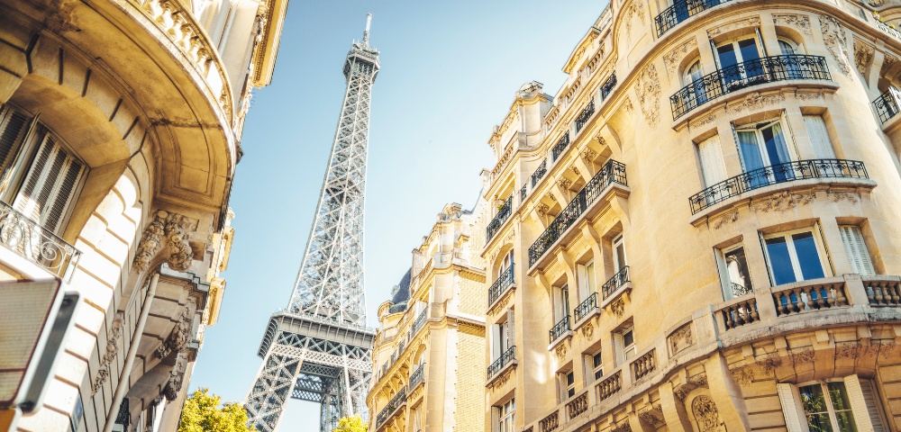 The Eiffel Tower rises between elegant Parisian buildings under a clear blue sky. Sunlight highlights the ornate facades, creating a warm, inviting atmosphere.