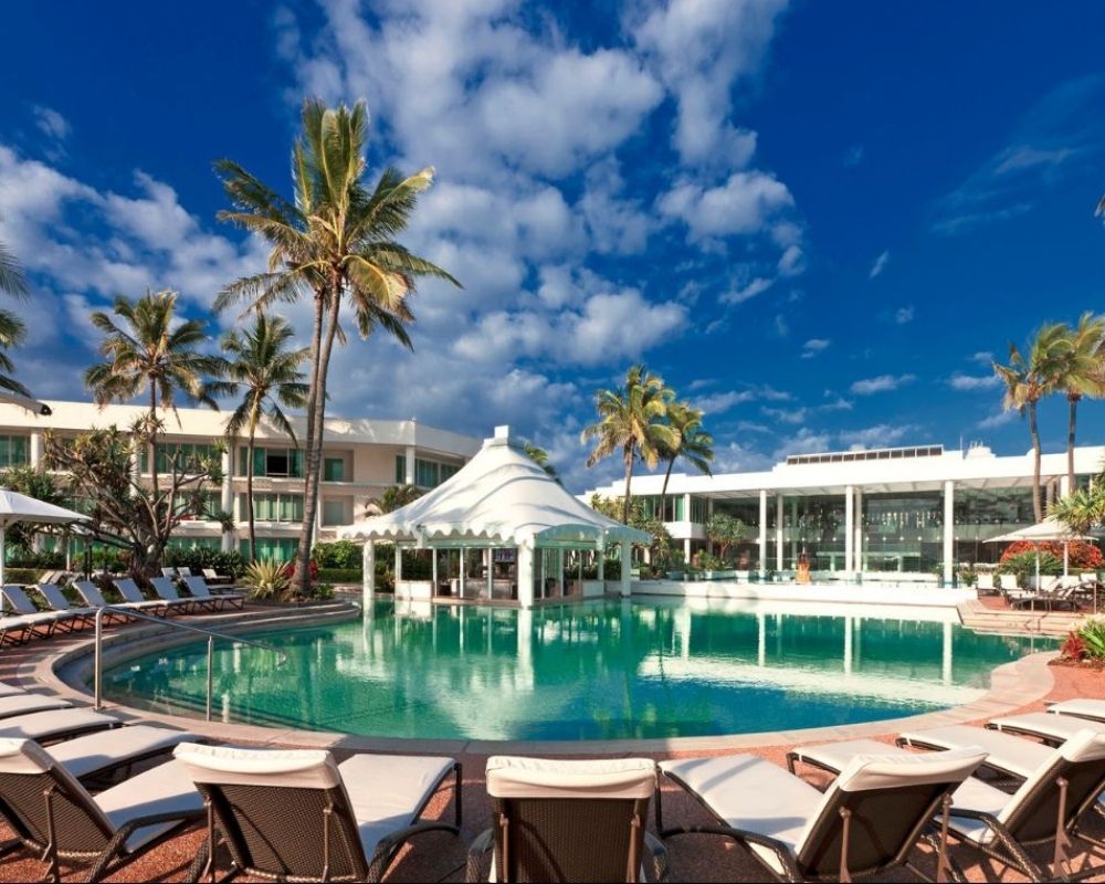 Luxurious resort pool area with lounge chairs and palm trees under a vibrant blue sky. A white canopy structure is central, evoking relaxation and elegance.