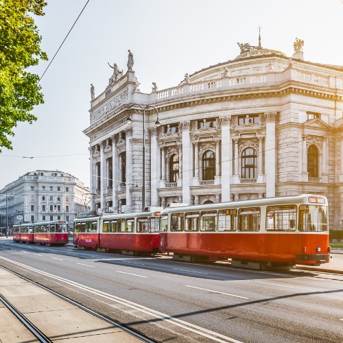 Red trams pass a grand, historic building with ornate columns under a bright sky, conveying a sense of classic European elegance and bustling city life.