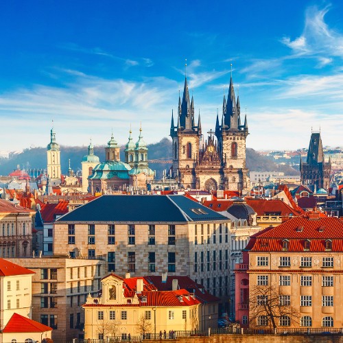 A vibrant cityscape of Prague with red-roofed buildings, Gothic spires of the Church of Our Lady before Týn, under a bright blue sky with wispy clouds.