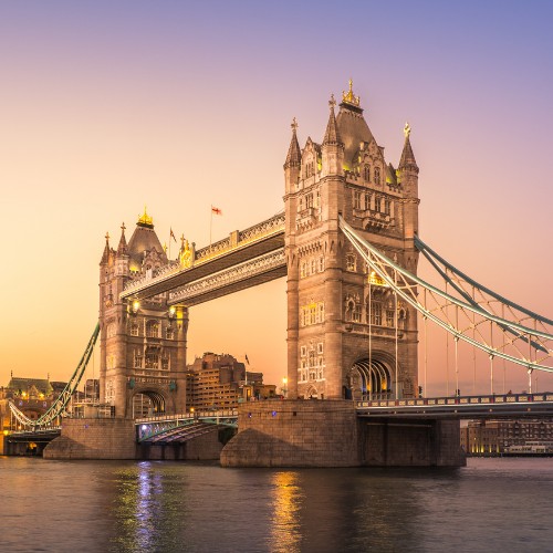Tower Bridge in London at sunset, illuminated with warm lights. The sky transitions from orange to purple, reflecting on the calm river below.