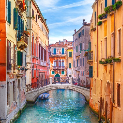 A picturesque Venice canal with pastel-colored buildings lining the water, a small arched bridge, and a blue sky. The scene feels serene and timeless.