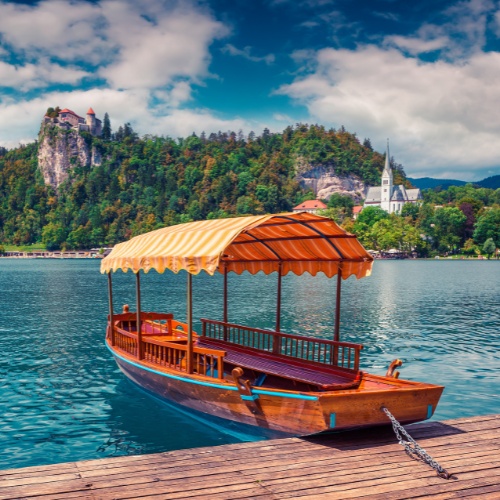 A wooden boat with a striped canopy is moored on a serene lake. A lush, tree-covered hill and a church with a steeple are in the background under a partly cloudy sky.