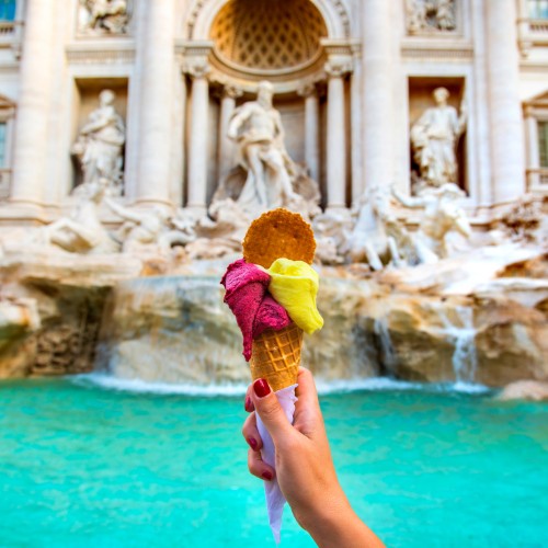 A hand holds a vibrant gelato cone in front of Rome's Trevi Fountain. The colorful ice cream contrasts with the historic marble statues and turquoise water.