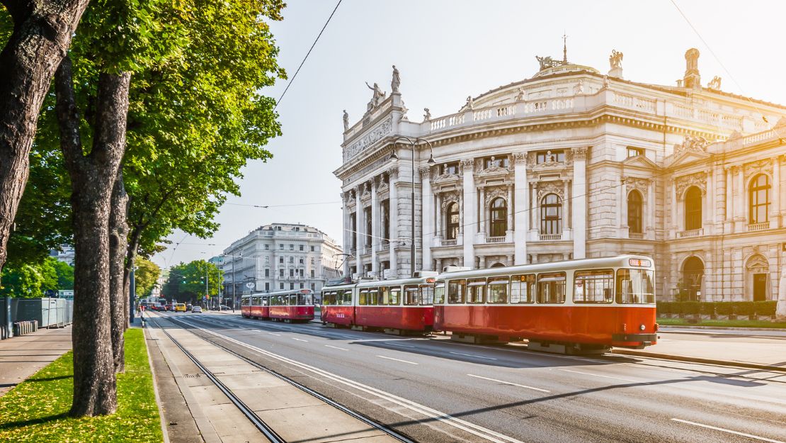 Red trams pass a grand, historic building with ornate columns under a bright sky, conveying a sense of classic European elegance and bustling city life.