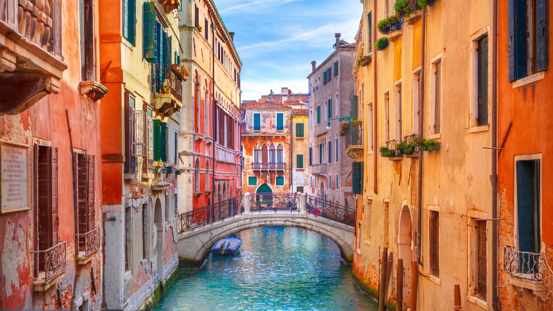A picturesque Venice canal with pastel-colored buildings lining the water, a small arched bridge, and a blue sky. The scene feels serene and timeless.
