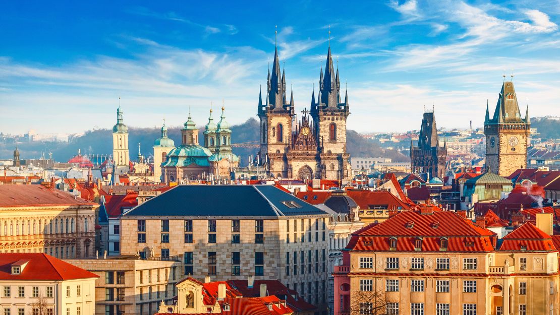 A vibrant cityscape of Prague with red-roofed buildings, Gothic spires of the Church of Our Lady before Týn, under a bright blue sky with wispy clouds.