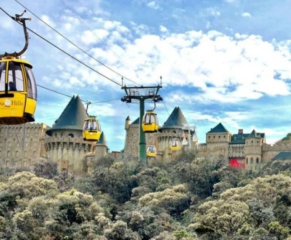Yellow cable cars ascend towards a European-style castle complex perched on a wooded hillside under a cloudy blue sky.