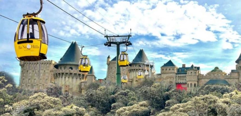 Yellow cable cars ascend towards a European-style castle complex perched on a wooded hillside under a cloudy blue sky.