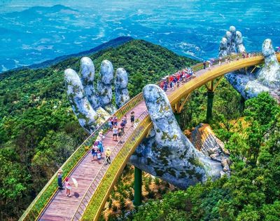 A golden bridge supported by two giant, moss-covered stone hands, curving over a lush, green mountainous landscape with a hazy blue sky in the distance. Many people walk along the bridge's pathways.