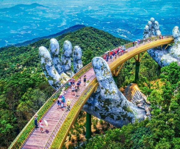 A golden bridge supported by two giant, moss-covered stone hands, curving over a lush, green mountainous landscape with a hazy blue sky in the distance. Many people walk along the bridge's pathways.