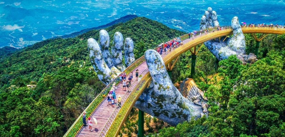 A golden bridge supported by two giant, moss-covered stone hands, curving over a lush, green mountainous landscape with a hazy blue sky in the distance. Many people walk along the bridge's pathways.