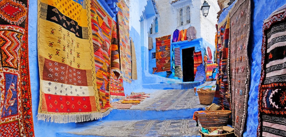 Vibrant, multicolored rugs hang along narrow, blue-painted alleyways. Baskets lie on cobblestone steps, evoking a lively, traditional market scene.