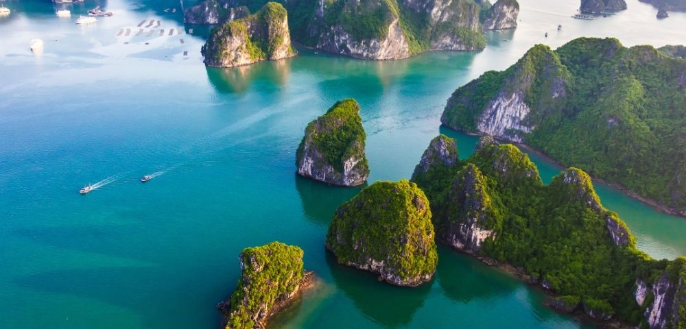 An aerial view of emerald green, jungle-covered limestone karst islands dotting turquoise waters, with two small boats sailing in the distance.