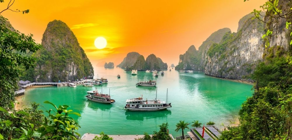 Boats navigate emerald green waters surrounded by lush, rocky karst islands under a vibrant orange sunset sky.