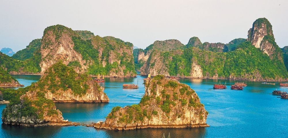 View of numerous karst islands covered in lush green vegetation rising from calm turquoise waters, with several traditional wooden boats sailing between them.