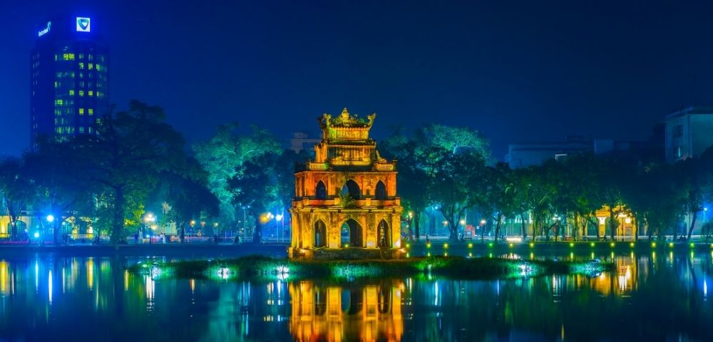 A multi-tiered pagoda-like structure is illuminated at night, situated on a small island in the middle of a calm body of water, with its warm, golden light reflecting on the surface.