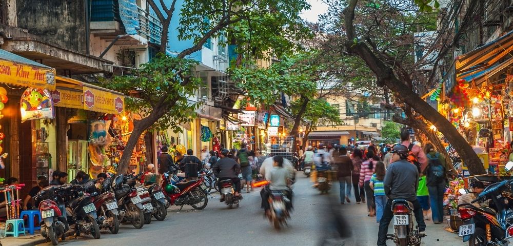 A busy street scene in a Southeast Asian city at dusk, with many motorbikes on the road and shops lining the sidewalks.
