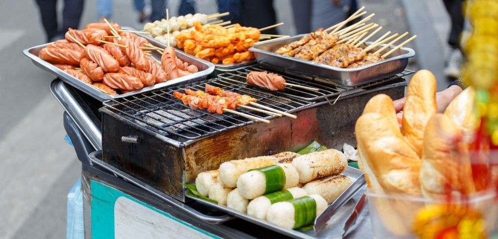 A street food cart grilling sausages and skewered meats, with rice cakes wrapped in leaves and loaves of bread on display.