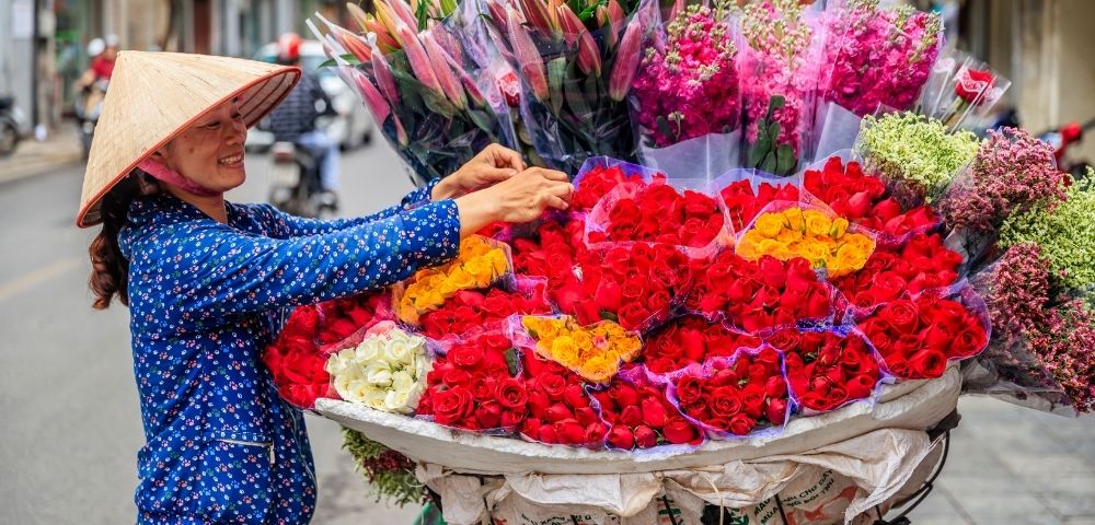 A street vendor wearing a conical hat arranges bouquets of red, yellow, and white roses on a bicycle cart overflowing with flowers.
