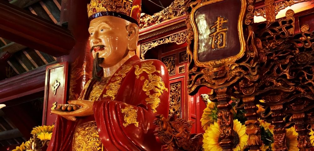 A statue of a man in ornate red and gold robes and a crown, with a long beard, stands within a richly decorated temple setting.