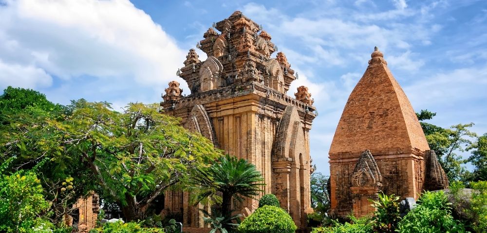 Ancient brick Cham towers with ornate tiered roofs stand amidst lush green trees and foliage under a bright blue sky with scattered clouds.