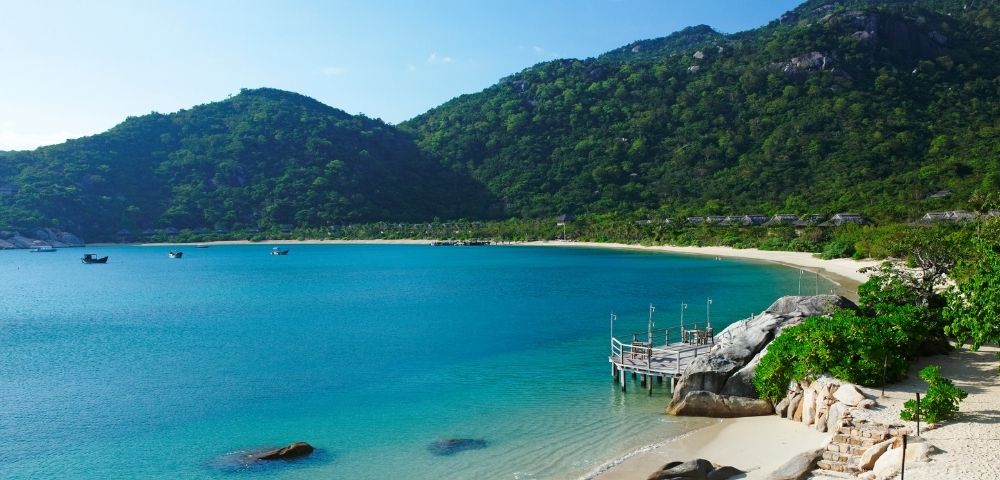 A serene tropical bay with calm, turquoise water, a sandy beach, a wooden pier extending into the sea, and lush green mountains in the background.
