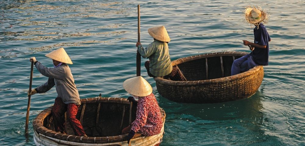 Four individuals in conical hats navigate the sea in traditional round fishing boats, with two rowing and two seated.