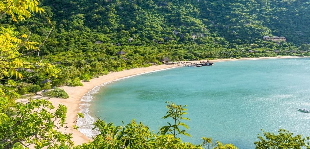 A crescent-shaped sandy beach borders a tranquil turquoise bay, backed by a lush green forested mountain with scattered buildings and a pier with several boats.