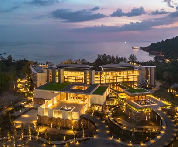 Aerial view of a modern, multi-story resort complex with illuminated windows and green roofs, nestled amongst lush trees and overlooking a calm ocean at dusk.