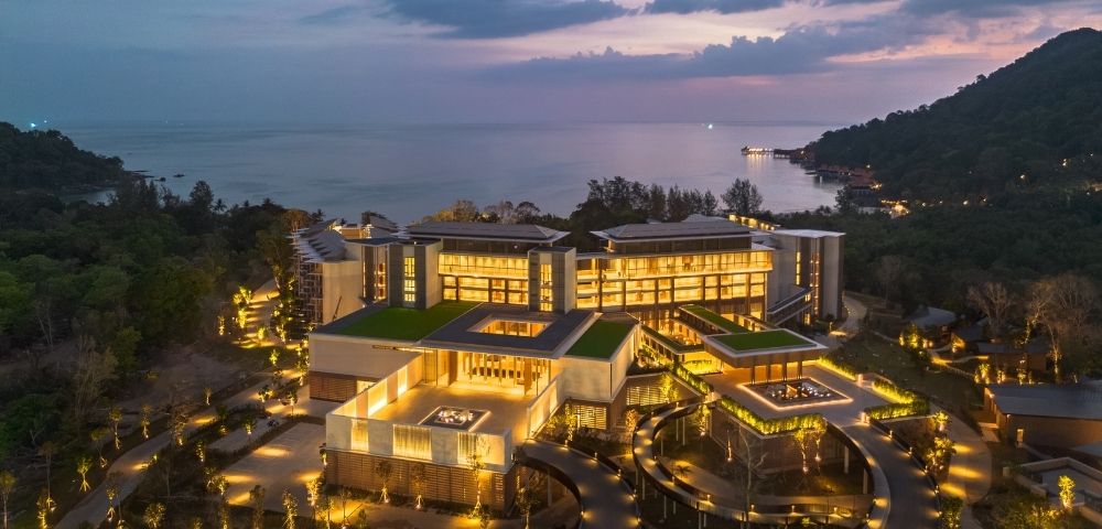 Aerial view of a modern, multi-story resort complex with illuminated windows and green roofs, nestled amongst lush trees and overlooking a calm ocean at dusk.