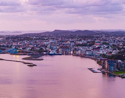 Panoramic view of a coastal city at twilight with a harbor, industrial buildings, and residential areas nestled against rolling hills under a soft purple sky.