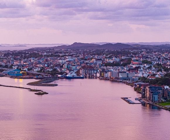 Panoramic view of a coastal city at twilight with a harbor, industrial buildings, and residential areas nestled against rolling hills under a soft purple sky.