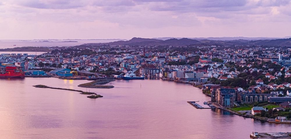 Panoramic view of a coastal city at twilight with a harbor, industrial buildings, and residential areas nestled against rolling hills under a soft purple sky.