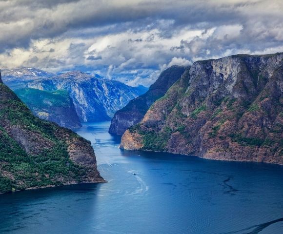 A wide, deep blue fjord carves through dramatic, steep, tree-covered mountains under a dramatic, cloudy sky.