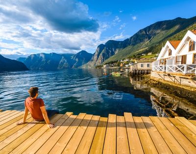 A person sits on a wooden pier, gazing out at a serene fjord with steep, verdant mountains and a quaint village on the shore.
