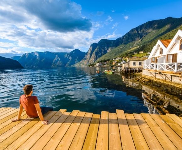 A person sits on a wooden pier, gazing out at a serene fjord with steep, verdant mountains and a quaint village on the shore.