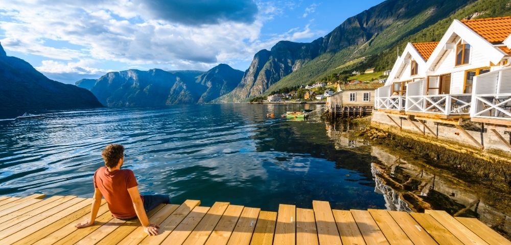 A person sits on a wooden pier, gazing out at a serene fjord with steep, verdant mountains and a quaint village on the shore.