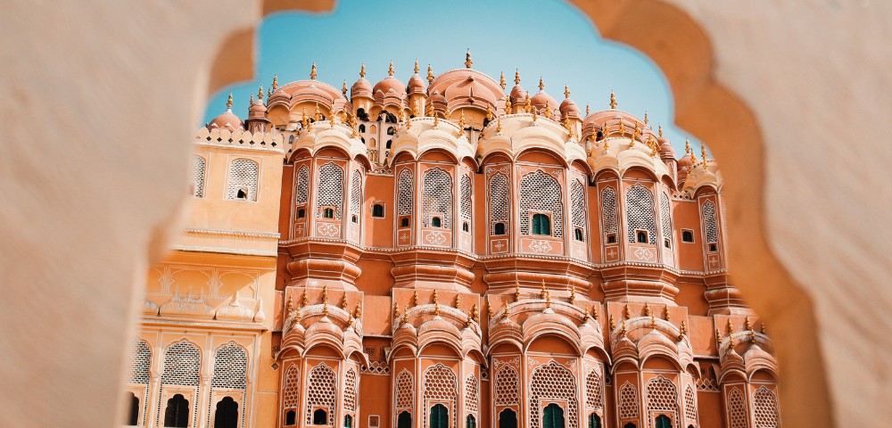 View of Hawa Mahal's ornate red and pink facade through an arched window, set against a clear blue sky, highlighting its intricate architecture.