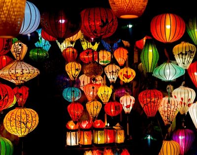 Colorful, illuminated paper lanterns of various shapes and sizes hanging closely together against a dark background.