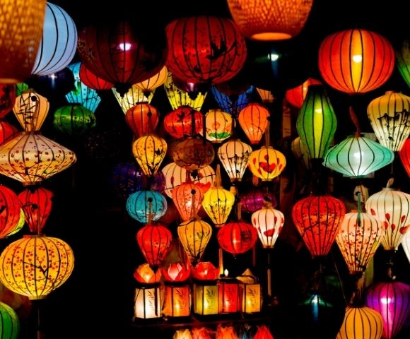Colorful, illuminated paper lanterns of various shapes and sizes hanging closely together against a dark background.