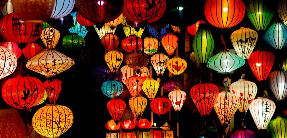 Colorful, illuminated paper lanterns of various shapes and sizes hanging closely together against a dark background.