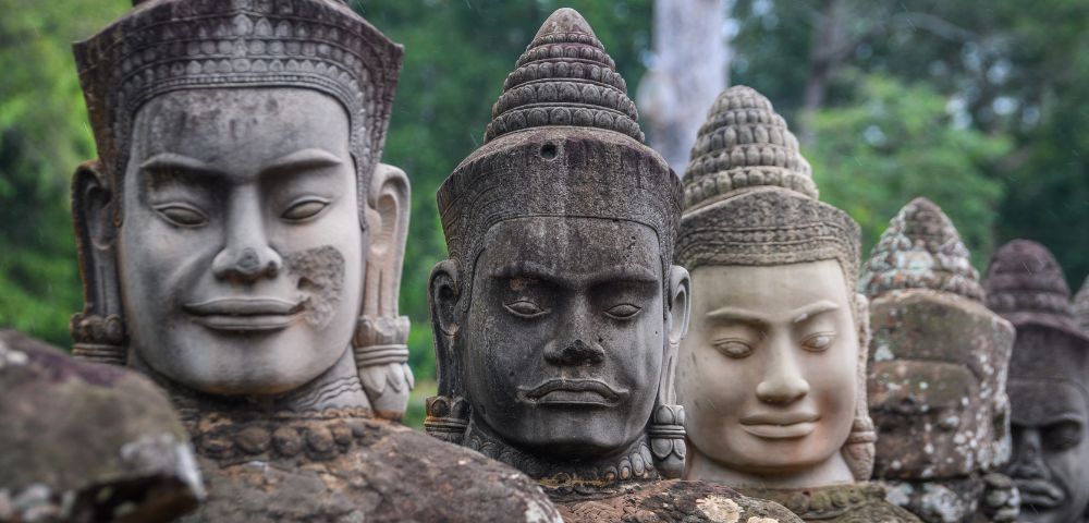 Close-up of ancient stone statues with serene expressions, featuring intricate headpieces and varied textures. Lush green forest in the background.
