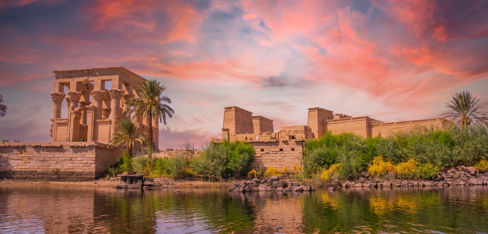 Ancient stone temple by a river, framed by palm trees and lush greenery. A vibrant sunset with pink and orange hues fills the sky, creating a serene and historic ambiance.